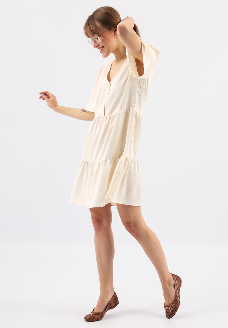 Cremefarbenes kurzes Kleid mit V-Ausschnitt, Knopfdetails, gestuftem Rüschendesign und kurzen Ärmeln; kombiniert mit braunen, perforierten Ballerinas.