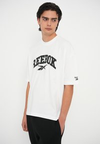 Bela bombažna majica s kratkimi rokavi, ki na prsih nosi velik črn logotip "REEBOK" in majhen logotip na rokavu. Ohlapen kroj in kratki rokavi.
