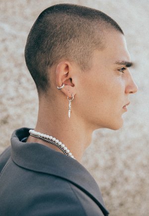 Silver earrings, including a knife-shaped charm, with a textured pearl necklace and a silver chain, worn with a gray collared jacket.