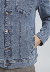 Denim jakna svijetloplave boje klasičnog kroja, sa dva džepa na prsima, kopčanjem na dugmad i vidljivim detaljima šavova.