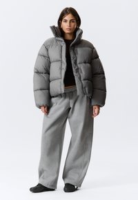 Sivá puffrová bunda s vysokým golierom, prešívaným vzorom a zipsom, nosená cez čierny top a sivé široké nohavice. Čierne nazúvacie topánky.