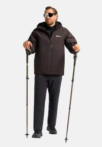 Veste imperméable noire avec capuche, portée sur un haut sombre et un pantalon noir robuste, tenant des bâtons de trekking avec des accents verts.