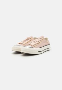 Light pink canvas sneakers with white rubber soles, gold eyelets, and black stripe accents. Low-top design with round toe.