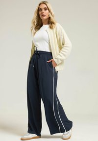 Pantalones de pierna ancha de color marino con franjas laterales blancas, cinturilla elástica y cordón. Combinados con una blusa blanca y un cardigans de punto crema.