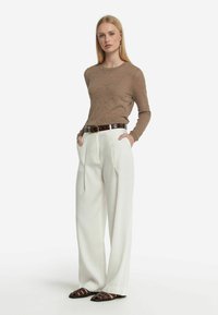 Maglione marrone con texture, pantaloni bianchi a pieghe larghi, cintura marrone scuro e sandali con cinturini marroni. Il modello sta in piedi con sicurezza con le mani nelle tasche.