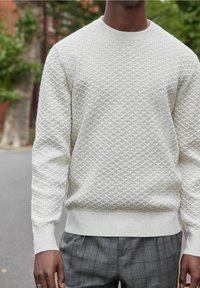 Pull en maille beige clair avec un motif de diamant texturé, des poignets et un ourlet côtelés. Associé à un pantalon à carreaux gris, montrant une coupe décontractée.