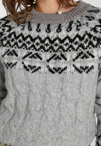 Grå stickad tröja med flätmönster, med ett svartvitt geometriskt motiv vid oket och en silverfärgad krage med pärlor.