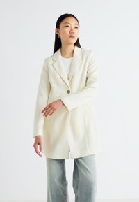 Blazer crema realizzato in tessuto morbido, con rever a punta, chiusura con un solo bottone e tasche laterali, abbinato a jeans in denim chiaro.