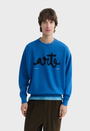 Junger Mann mit mittellangen braunen Haaren, der einen blauen Pullover mit dem Schriftzug "arte" in schwarzer Handschrift trägt, darüber ein hellblaues Hemd und braune Hose.