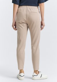 Pantalones elásticos beige con un ajuste slim, cinturilla elástica y un bolsillo trasero, combinados con zapatillas blancas. Textura suave y diseño a medida.