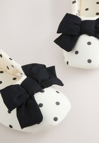 Chaussures bébé crème à pois noirs avec de grands nœuds en tissu noir dessus, présentées sur un fond clair.