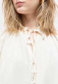 Κοντινή λήψη του λαιμού και των ώμων μιας γυναίκας που φοράει λευκό πουκάμισο με δεμένους φιόγκους στον γιακά και ατημέλητα, υφασμάτινα ξανθά μαλλιά.