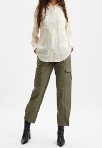 Camicia abbottonata color pesca chiaro con due tasche sul petto, abbinata a pantaloni cargo verde oliva. Stivali neri con tacco completano l'outfit.