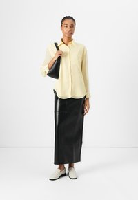 Camicetta gialla abbottonata, gonna in pelle nera e scarpe slip-on di colore chiaro. La borsa è piccola e nera, con una forma strutturata.