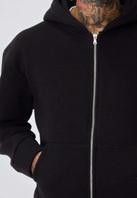 Personne portant un sweat à capuche noir zippé, les mains dans les poches avant, montrant des tatouages sur le cou et le poignet.