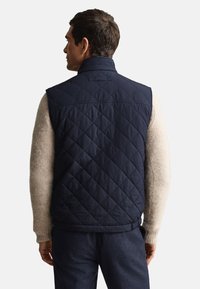 Marinblå quiltad väst av syntetiskt tyg, med hög krage och elastisk midjeband, bärs över en beige tröja och visar detaljerad sömnad.