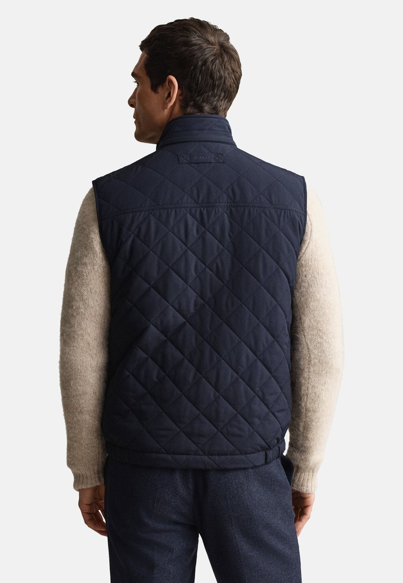 Chaleco acolchado azul marino hecho de tela sintética, con cuello alto y cinturilla elástica, llevado sobre un suéter beige, mostrando costuras detalladas.