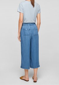 Woman wearing light blue cropped wide-leg jeans with buttoned back pockets, light grey t-shirt, and brown woven slip-on sandals, standing.