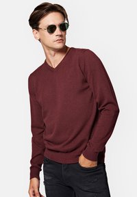 Burgundowy sweter w serek wykonany z miękkiego materiału, w połączeniu z zielonymi okularami przeciwsłonecznymi, noszony z przetartymi czarnymi dżinsami.