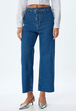 Jeans en denim bleu taille haute avec une coupe large, présentant un ourlet effrangé et une décoloration subtile. Associés à des escarpins argentés.