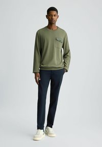 Maglietta a maniche lunghe verde oliva con taschino sul petto, abbinata a pantaloni blu navy e sneakers bianche. Design semplice con vestibilità rilassata.