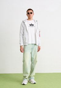 Grå hættetrøje, hvid t-shirt med sort logo, lysegrønne jeans og hvide sneakers. Runde sorte solbriller og minimale accessories.