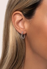 Pendiente de aro de plata con un diseño retorcido, que presenta pequeñas cuentas redondas a lo largo de la curva. Montado en la oreja de una persona, con pelo castaño claro cerca.