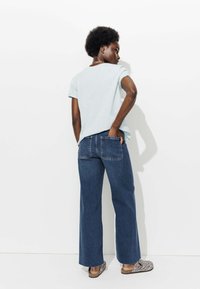 Chemise bleu clair à manches courtes associée à un jean tailles larges bleu foncé, avec des poches arrière. Les chaussures sont des sabots à motifs.