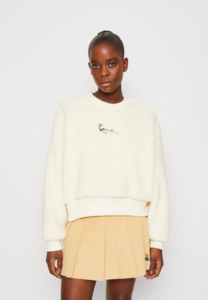 Kremfarget fleecegenser med rund hals, senkede skuldre og svart broderi. Kombinert med en plissert skjørtekjole i beige.