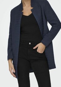 Cappotto lungo blu navy a motivo a spina di pesce con apertura frontale, colletto alla mandarino e maniche ampie, abbinato a jeans neri attillati.