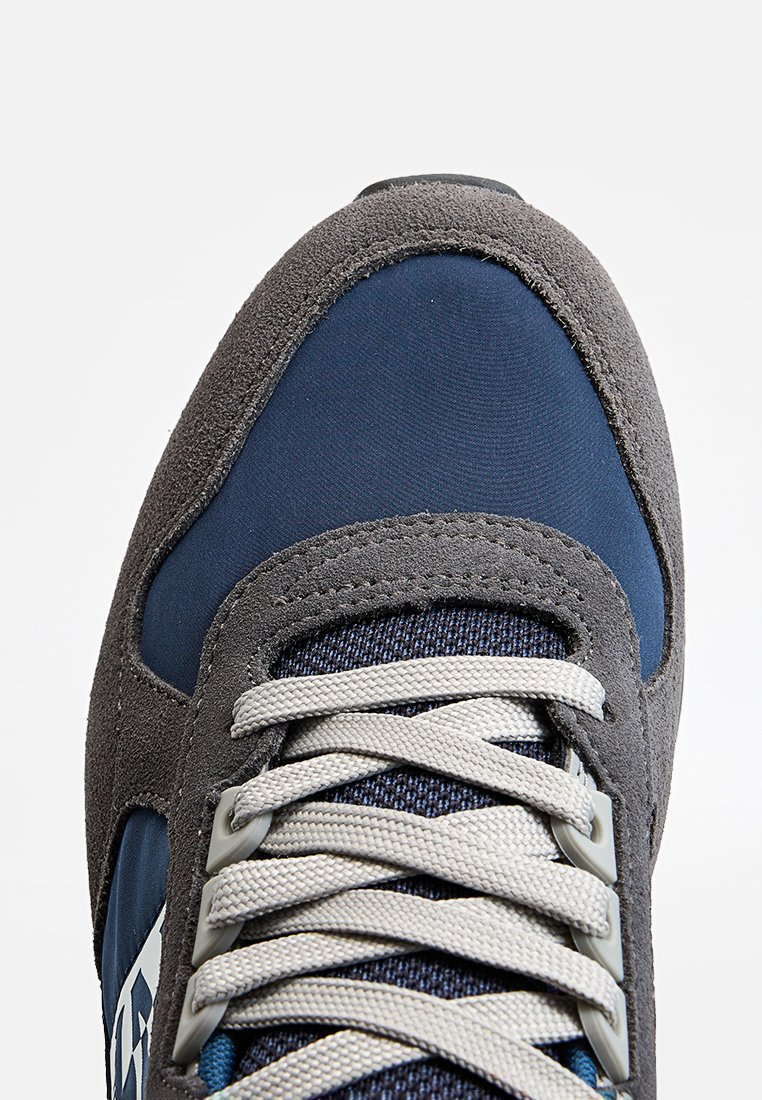Męski but sportowy z szaro-suede'owym wierzchem, granatowymi panelami tekstylnymi, szarymi sznurówkami i niebieskimi akcentami na języku. Widoczne szczegóły teksturowej powierzchni.