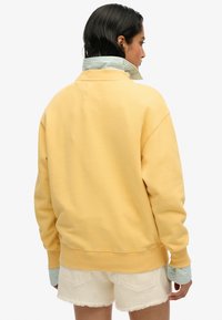 Sweat-shirt jaune à coupe décontractée, avec des poignets côtelés, un ourlet arrondi, et un col rayé bleu et blanc dépassant en dessous.