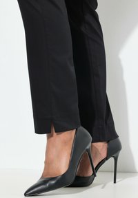 Schwarze, tapered Hosen mit einem Schlitz am Saum, kombiniert mit spitzen, schwarzen High Heels mit dünnem Absatz und glattem Ledertextur.