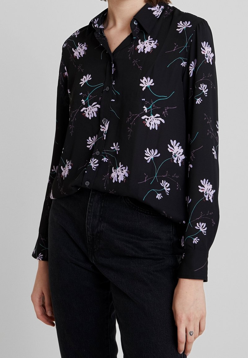 Femme portant un chemisier noir à boutons avec un motif floral violet et un jean noir, main avec un petit tatouage en forme de cœur visible sur le côté.