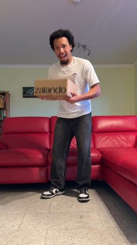 Scatola di cartone con "zalando" stampato in nero sul fronte. Persona che tiene la scatola, vestita con una camicia bianca e jeans scuri, su un divano rosso.