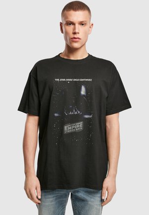 Jeune homme portant un t-shirt noir Star Wars The Empire Strikes Back avec un jean bleu, debout devant un fond uni gris clair.
