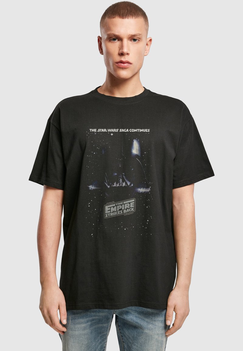 Jeune homme portant un t-shirt noir Star Wars The Empire Strikes Back avec un jean bleu, debout devant un fond uni gris clair.