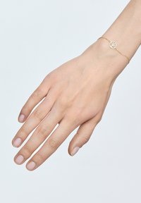 Złota bransoletka z okrągłym wisiorkiem z misternie wyciętym wzorem drzewa, noszona na ręce z naturalnymi paznokciami na jasnym tle.