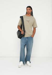 Camiseta de manga corta verde oliva claro con un gráfico impreso; combinada con unos vaqueros de denim azul de corte holgado y zapatillas blancas, llevando un bolso negro.