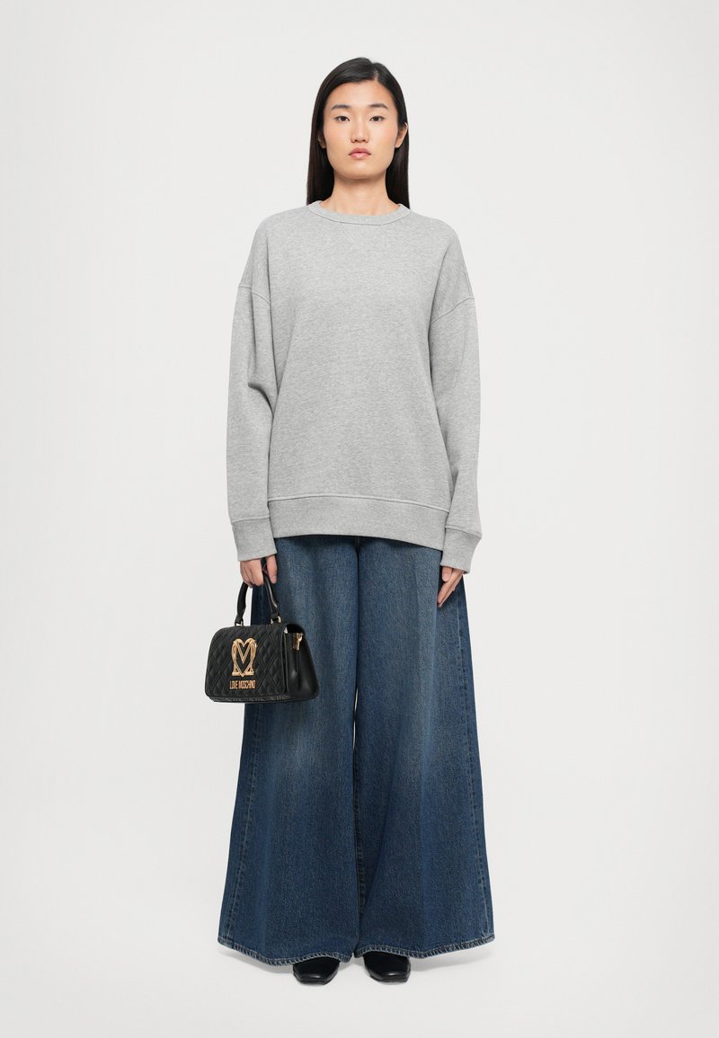 Frau trägt einen grauen, oversized Pullover und weit ausgestellte blaue Jeans. Sie hält eine schwarze, gesteppte Handtasche mit goldenen Logos und Griffen.