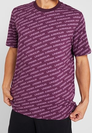 Camiseta de manga corta morada con cuello redondo, que presenta un patrón de texto "UNDER ARMOUR" repetido en tonos más claros. Textura de tejido suave.