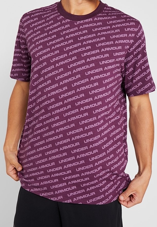Lila kortärmad t-shirt med rund hals, med ett upprepat "UNDER ARMOUR" textmönster i ljusare nyanser. Mjuk tygkvalitet.