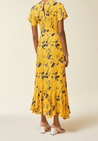 Żółta sukienka midi w kwiaty z falbanami na rękawach i otworem na plecach. Posiada falbanasty dół i fioletowy wzór kwiatowy. Noszona z białymi sandałami na słupku.