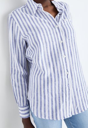 Kobieta ubrana w niebiesko-białą koszulę zapinaną na guziki w pionowe paski z długimi rękawami oraz jasnoniebieskie dżinsy, na białym tle.