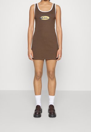 Kobieta ubrana w brązową sukienkę mini bez rękawów z białymi wykończeniami i żółto-czarnym logo na piersi, w połączeniu z białymi skarpetkami i brązowymi mokasynami.