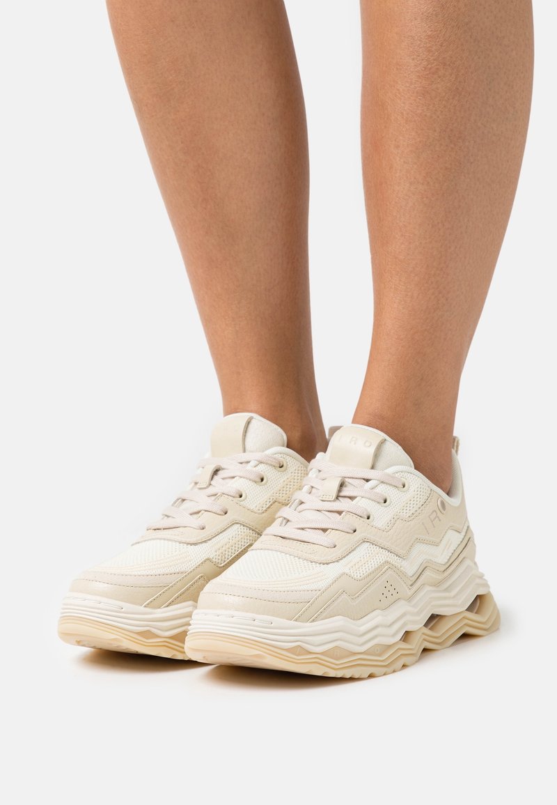Baskets beiges et blanches à lacets avec semelles à motif zigzag portées pieds nus sur fond blanc.
