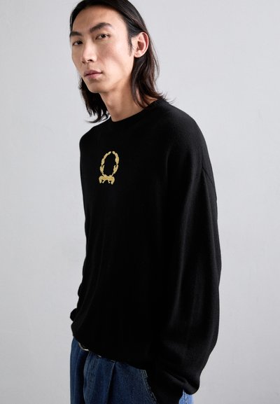 Schwarzer Pullover aus weichem Material, mit einem goldenen gestickten Logo mit Lorbeerkranz und Schleife auf der Brust. Legere Passform, lange Ärmel.