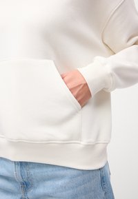 Felpa color crema con tasca frontale, polsini e orlo a coste; texture morbida, indossata con jeans azzurri chiari.