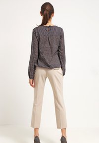 Blusa a pois grigio scuro con maniche lunghe e apertura a goccia sul retro, abbinata a pantaloni beige chiaro su misura e scarpe scure.
