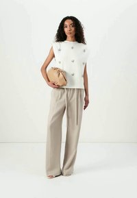Suéter em tecido branco com detalhes decorativos, combinado com calças largas em bege. Modelo segura uma clutch de bolsa em bege.